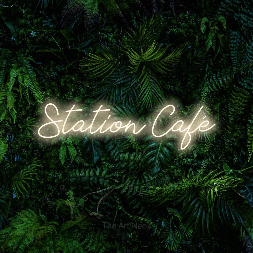 “Station café” LED neon sign in warm white, with cursive lettering, shown against dense tropical foliage.