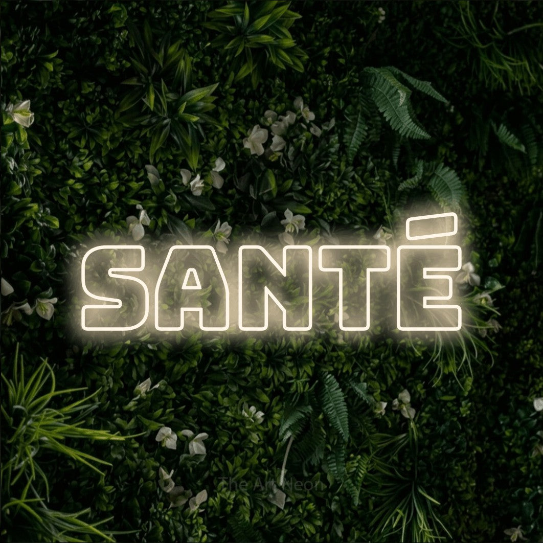 “Santé” LED neon sign in warm white, with bold outline lettering, shown against a dense green plant wall.
