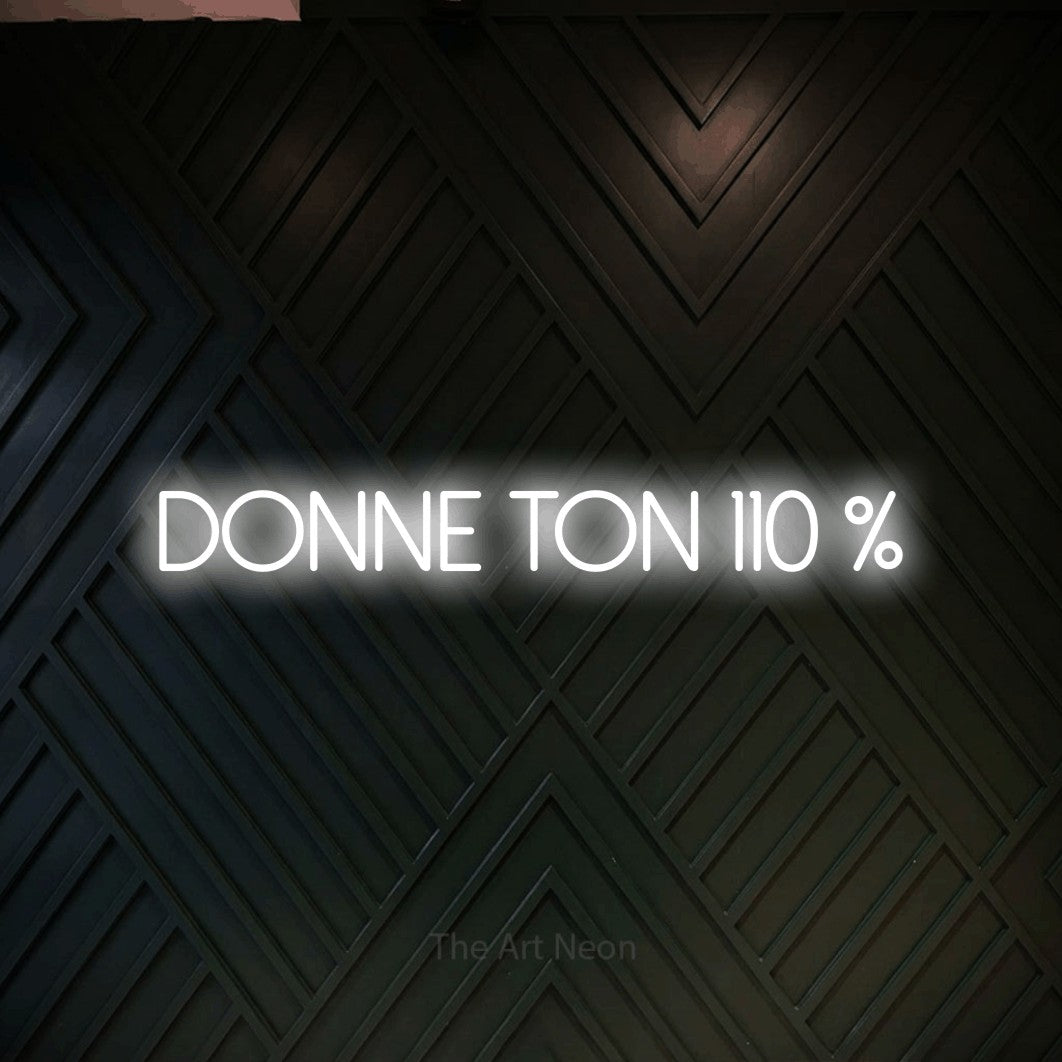 “Donne ton 110 %” LED neon sign in cool white, with thin sans-serif lettering, shown against a black chevron-patterned wood wall.