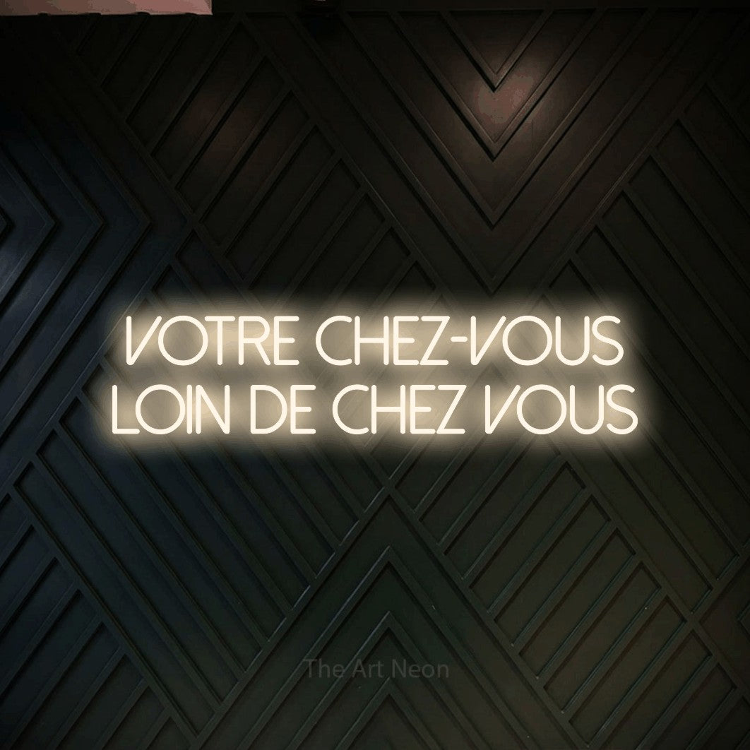 Lettrage en néon LED « Votre chez-vous loin de chez vous » sur deux lignes en blanc chaud, en lettres fines sans empattement, présenté sur un mur en bois noir à motif chevron, pour un hôtel.