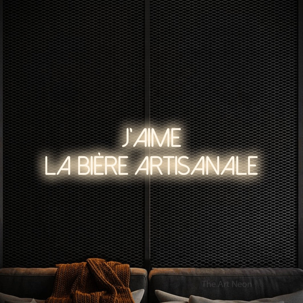 Composition en néon LED « J'aime la bière artisanale » sur deux lignes en blanc chaud, en lettres fines sans empattement, mise en valeur dans un coin salon moderne aux tons noirs, pour une microbrasserie.
