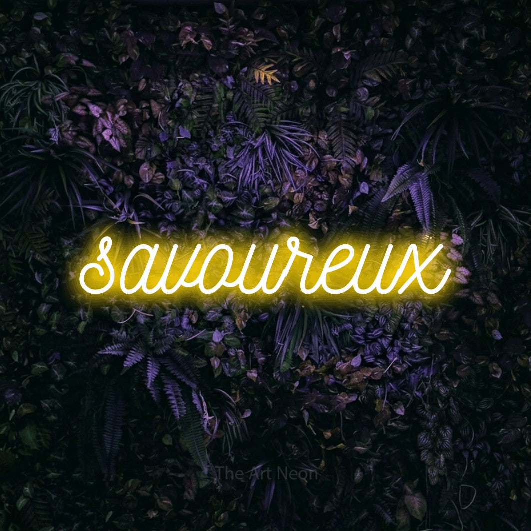 “Savoureux” LED neon sign in yellow, with ornate cursive lettering, shown against a dark wall with purple flowers.