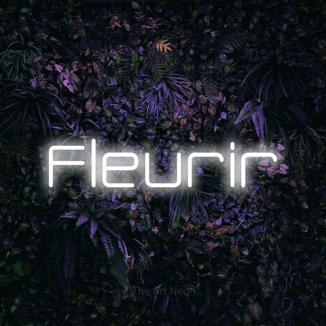 “Fleurir” LED neon sign in cool white, with rounded geometric lettering, shown against a dark wall with purple flowers.