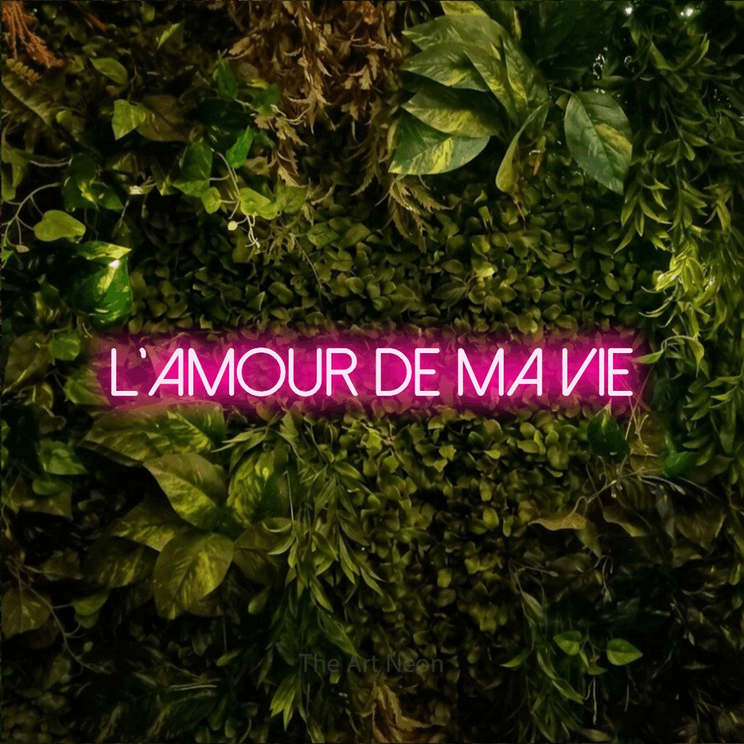 “L'amour de ma vie” LED neon sign in hot pink, with thin sans-serif lettering, shown against bright green and yellow foliage.