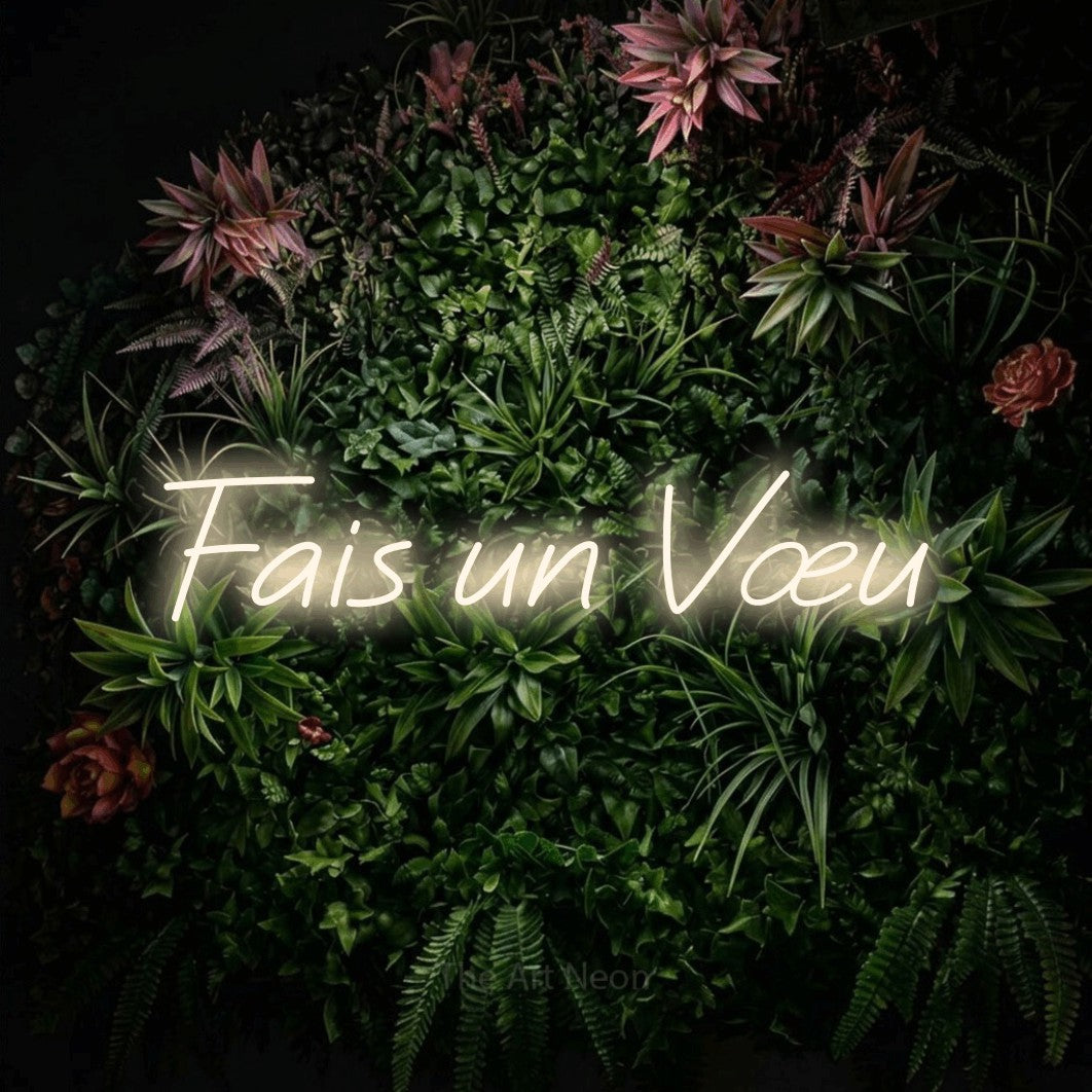“Fais un vœu” LED neon sign in warm white, with slanted cursive lettering, shown against a flowering plant wall.
