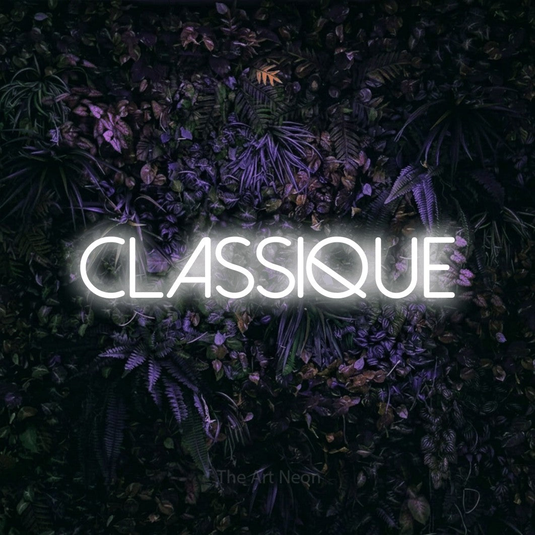 “Classique” LED neon sign in cool white, with thin sans-serif lettering, shown against a dark wall with purple flowers.