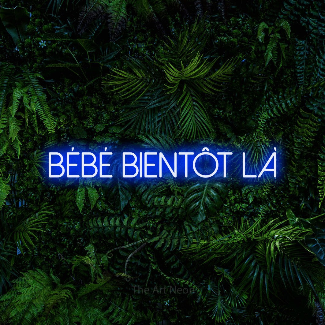 Composition en néon LED « Bébé bientôt là » en bleu, en lettres fines sans empattement, mise en valeur devant un feuillage tropical dense, pour une fête prénatale.