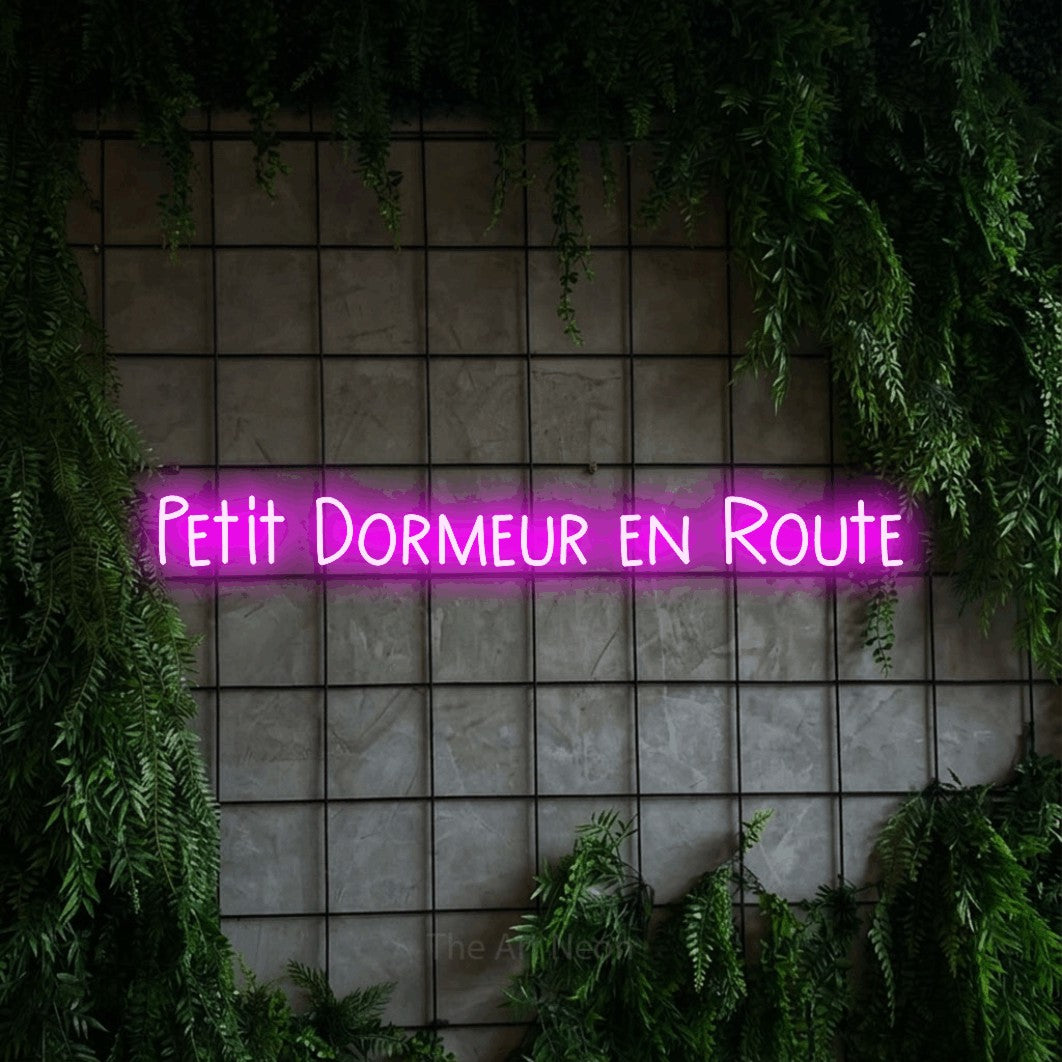 “Petit dormeur en route” LED neon sign in pink, with narrow hand-drawn lettering, shown against an ivy-covered concrete grid.