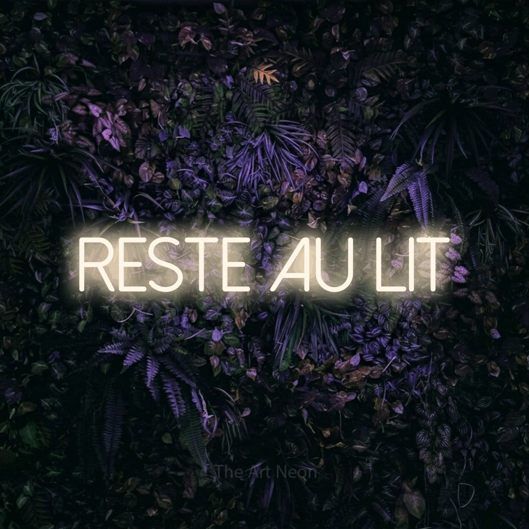 “Reste au lit” LED neon sign in warm white, with thin sans-serif lettering, shown against a dark wall with purple flowers.