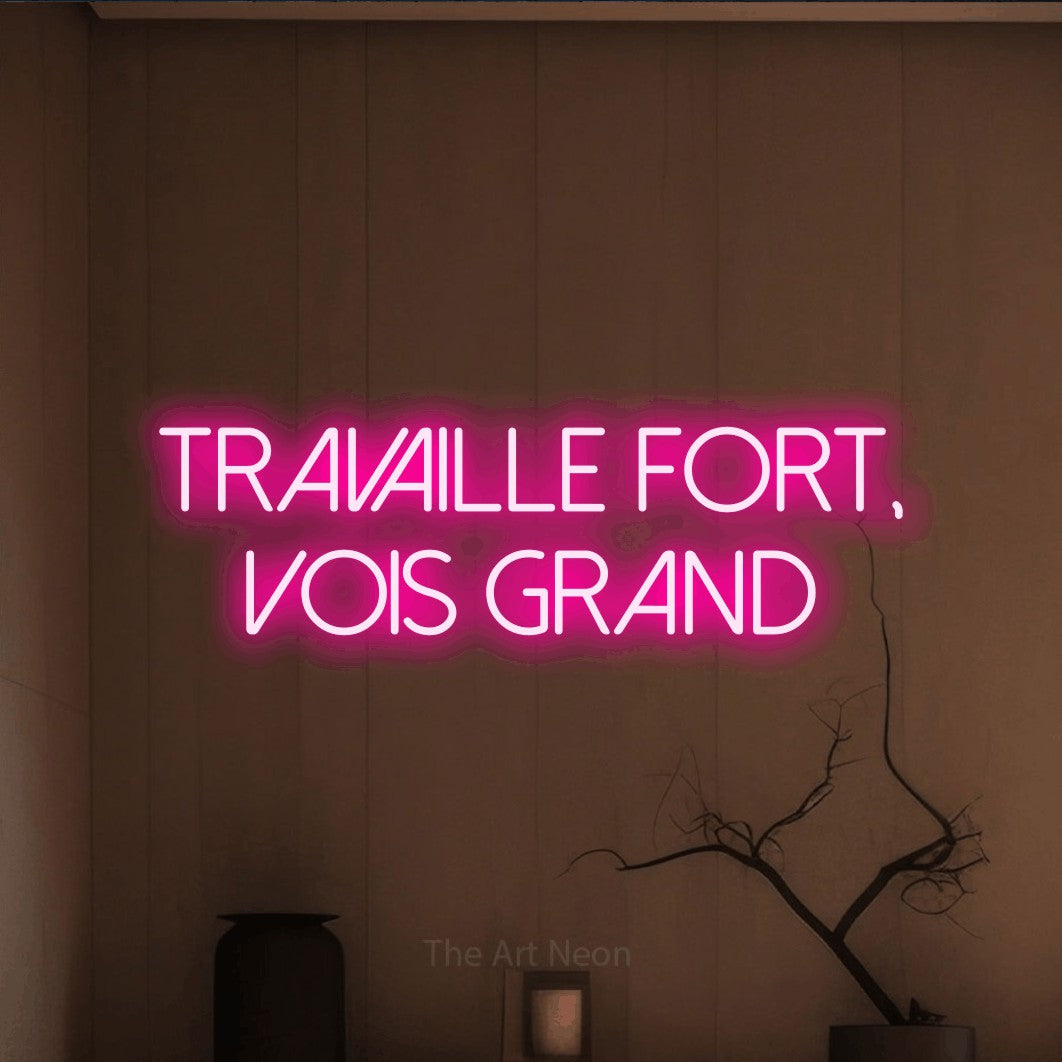 “Travaille fort, vois grand” LED neon sign in hot pink, with thin sans-serif lettering across 2 lines, shown against a simple earth-toned interior.