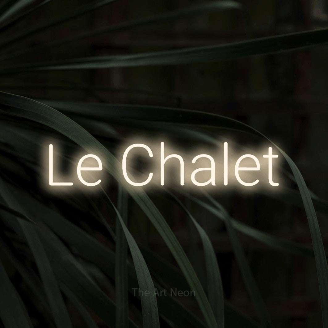 “Le chalet” LED neon sign in warm white, with rounded sans-serif lettering, shown against a close-up of dark foliage.