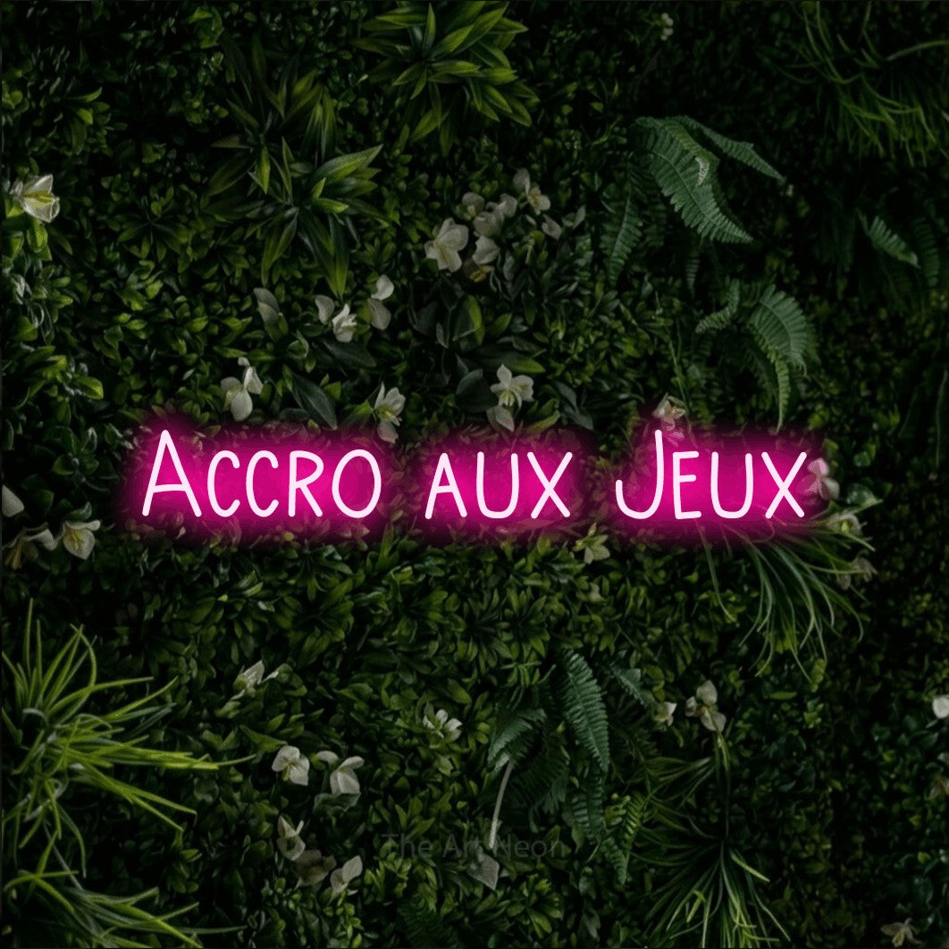 “Accro aux jeux” LED neon sign in hot pink, with narrow hand-drawn lettering, shown against a dense green plant wall.