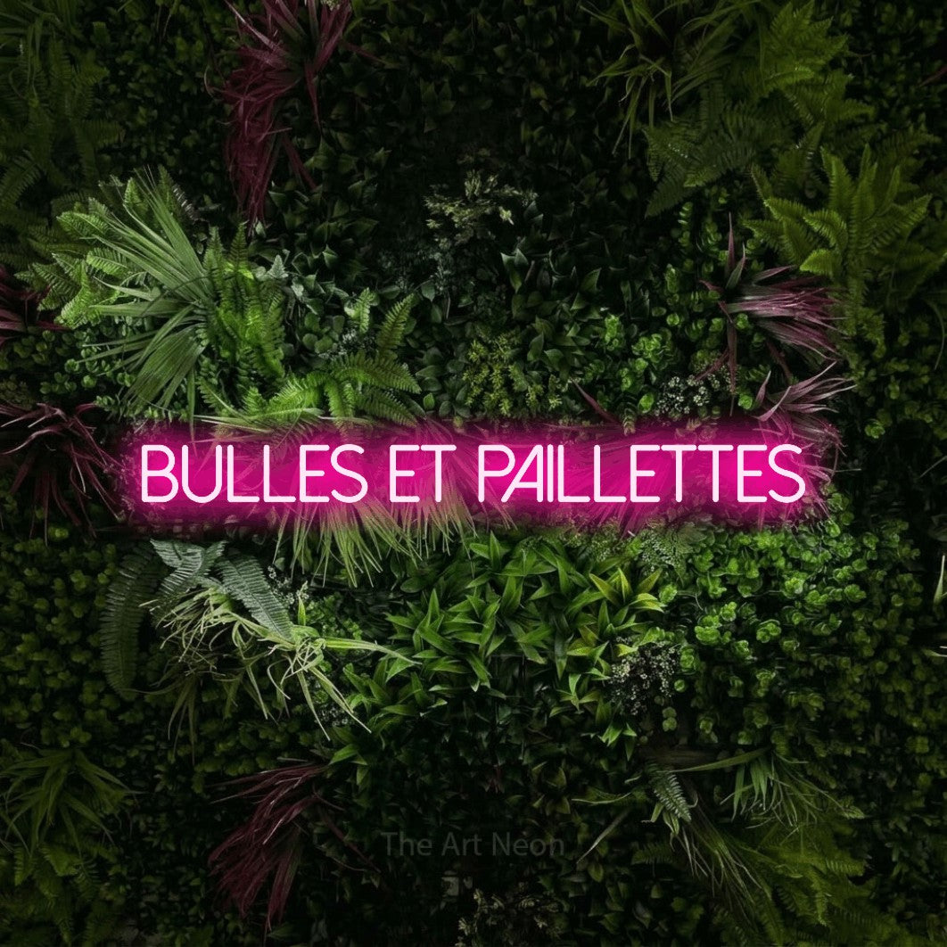 “Bulles et paillettes” LED neon sign in hot pink, with thin sans-serif lettering, shown against a plant wall with pink flowers.