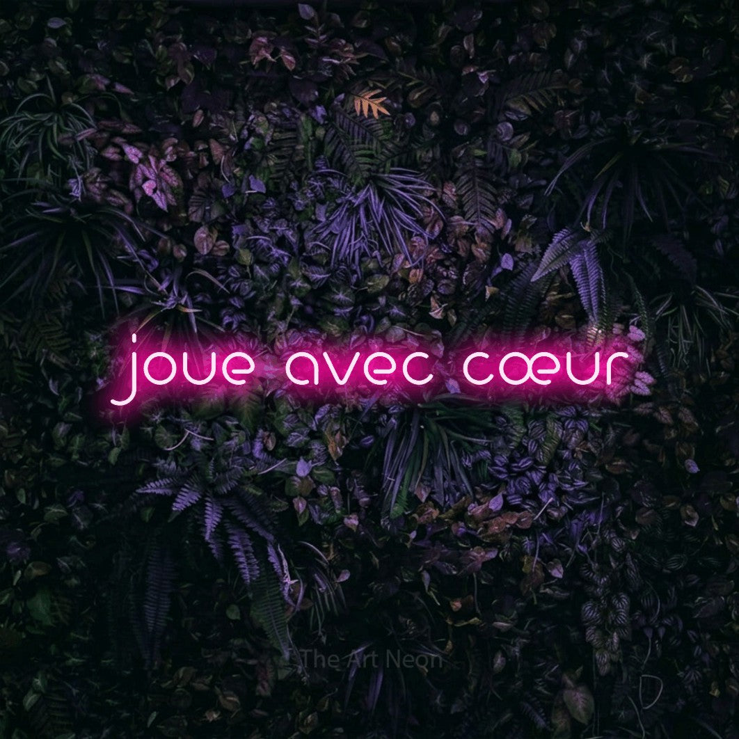 “Joue avec cœur” LED neon sign in hot pink, with stylised geometric lettering, shown against a dark wall with purple flowers.
