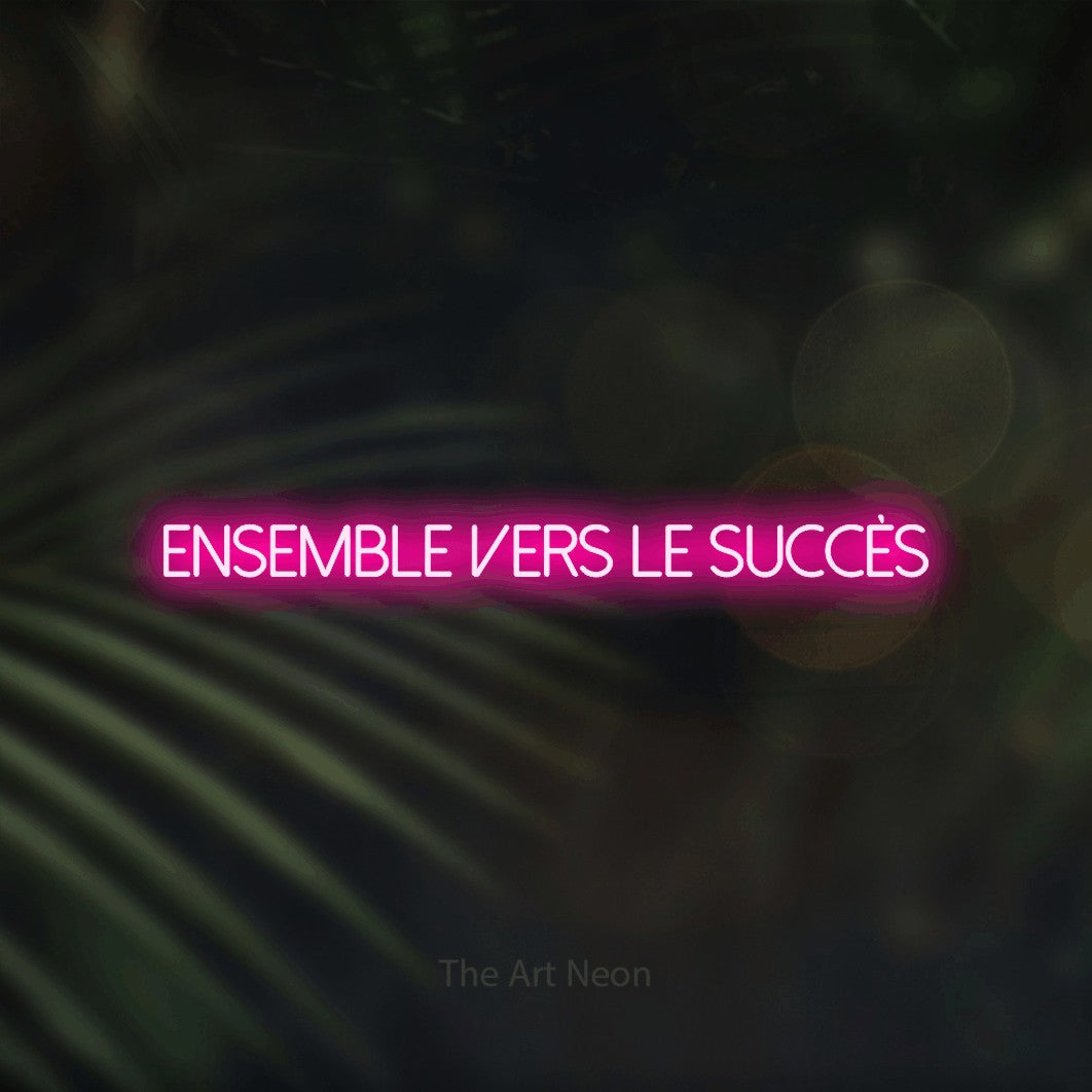 “Ensemble vers le succès” LED neon sign in hot pink, with thin sans-serif lettering, shown against a blurred palm and bokeh-light backdrop.