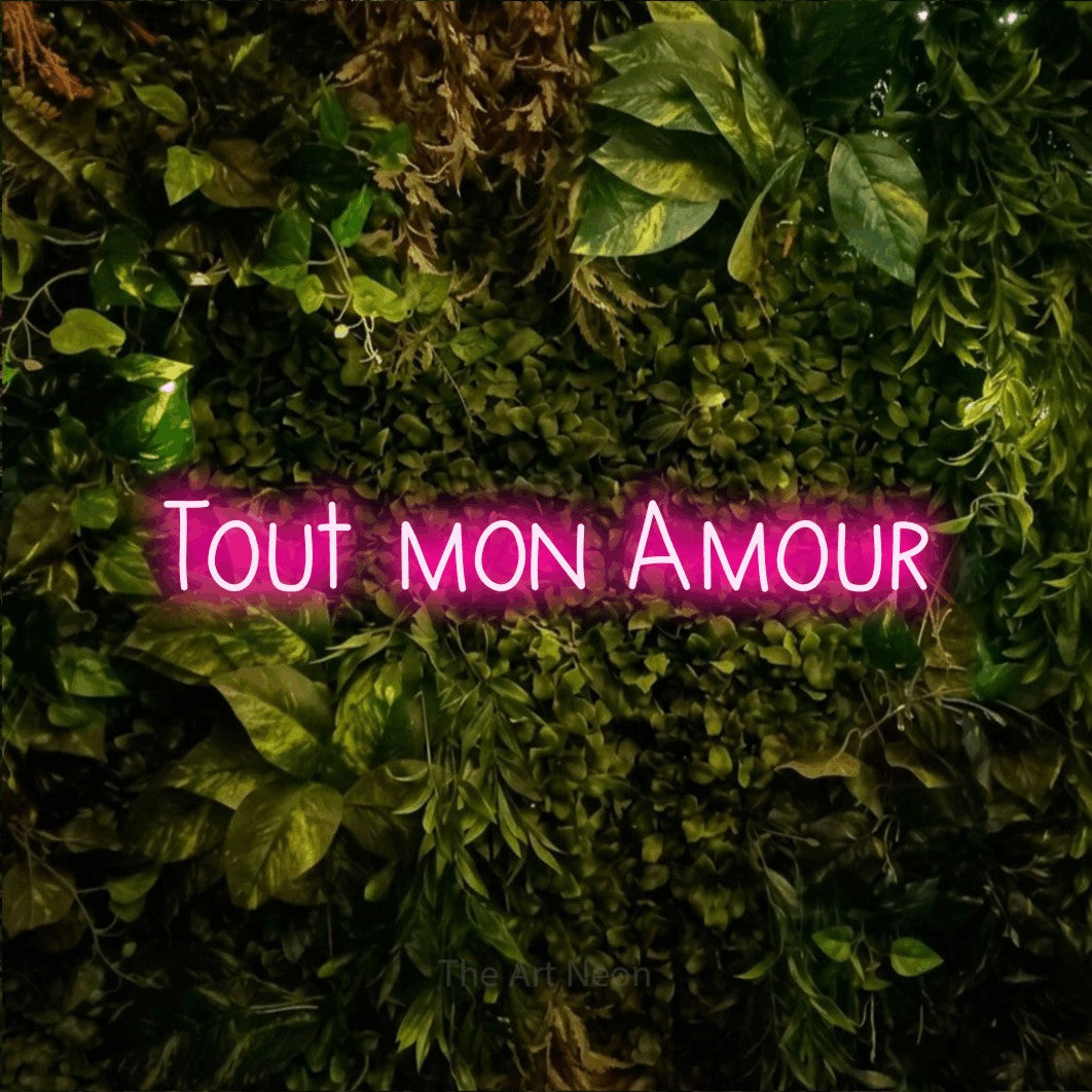 “Tout mon amour” LED neon sign in hot pink, with narrow hand-drawn lettering, shown against bright green and yellow foliage.