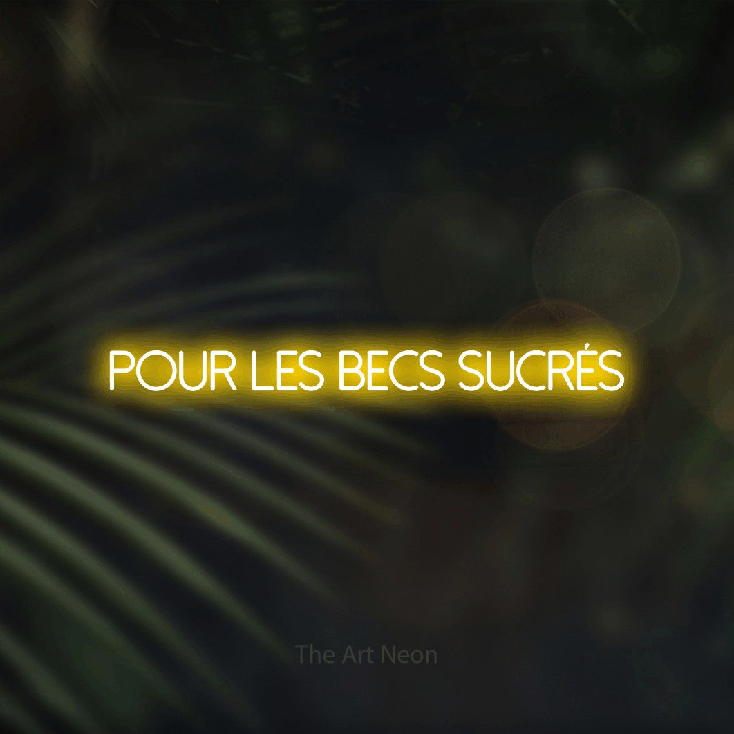“Pour les becs sucrés” LED neon sign in yellow, with thin sans-serif lettering, shown against a blurred palm and bokeh-light backdrop.