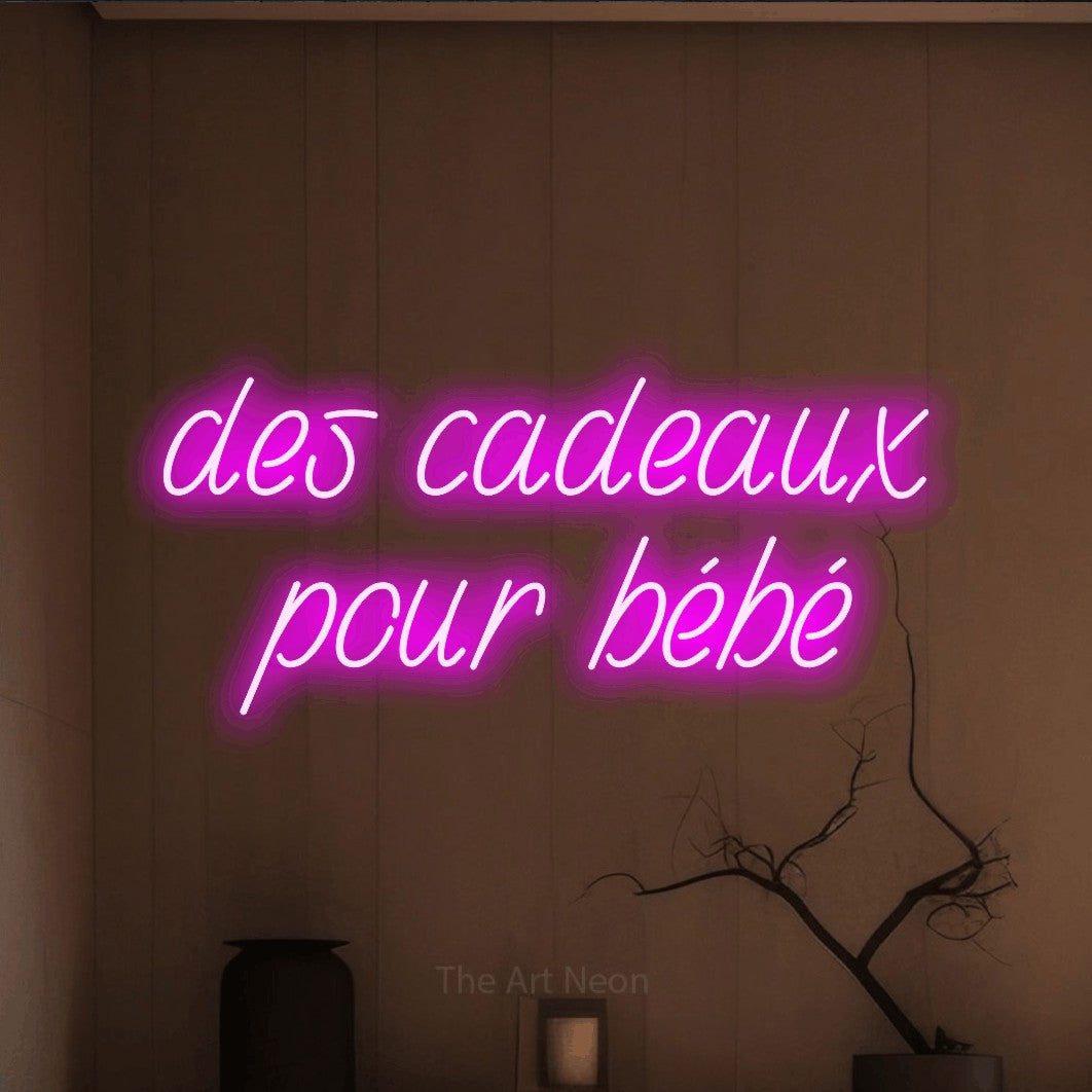 Composition en néon LED « Des cadeaux pour bébé » sur deux lignes en rose, en écriture manuscrite inclinée, mise en valeur dans un intérieur épuré aux tons terre, pour une fête prénatale.
