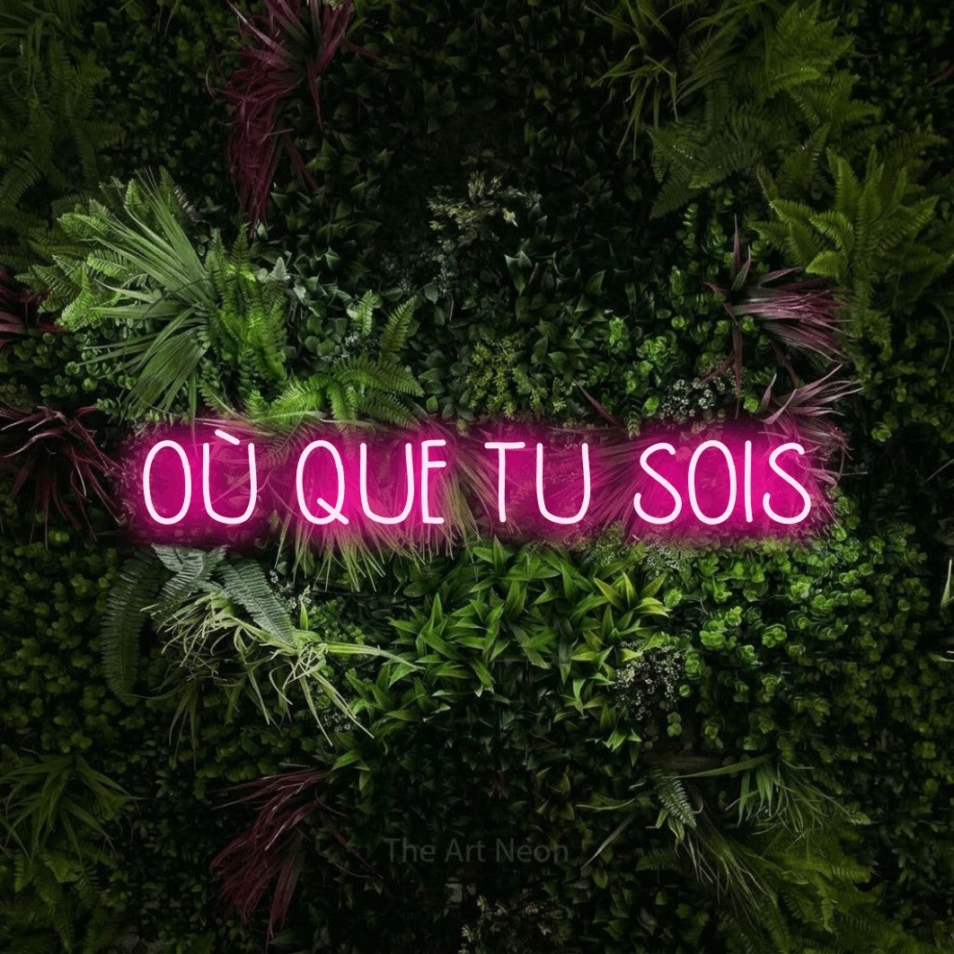 “Où que tu sois” LED neon sign in hot pink, with narrow hand-drawn lettering, shown against a plant wall with pink flowers.