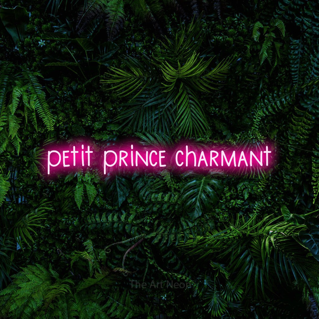 “Petit prince charmant” LED neon sign in hot pink, with narrow hand-drawn lettering, shown against dense tropical foliage.