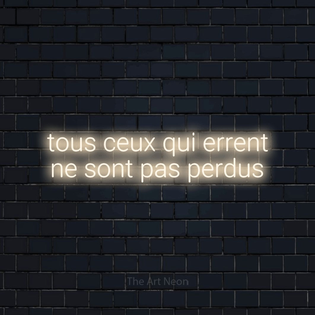 “Tous ceux qui errent ne sont pas perdus” LED neon sign in warm white, with rounded sans-serif lettering across 2 lines, shown against a dark navy brick wall.