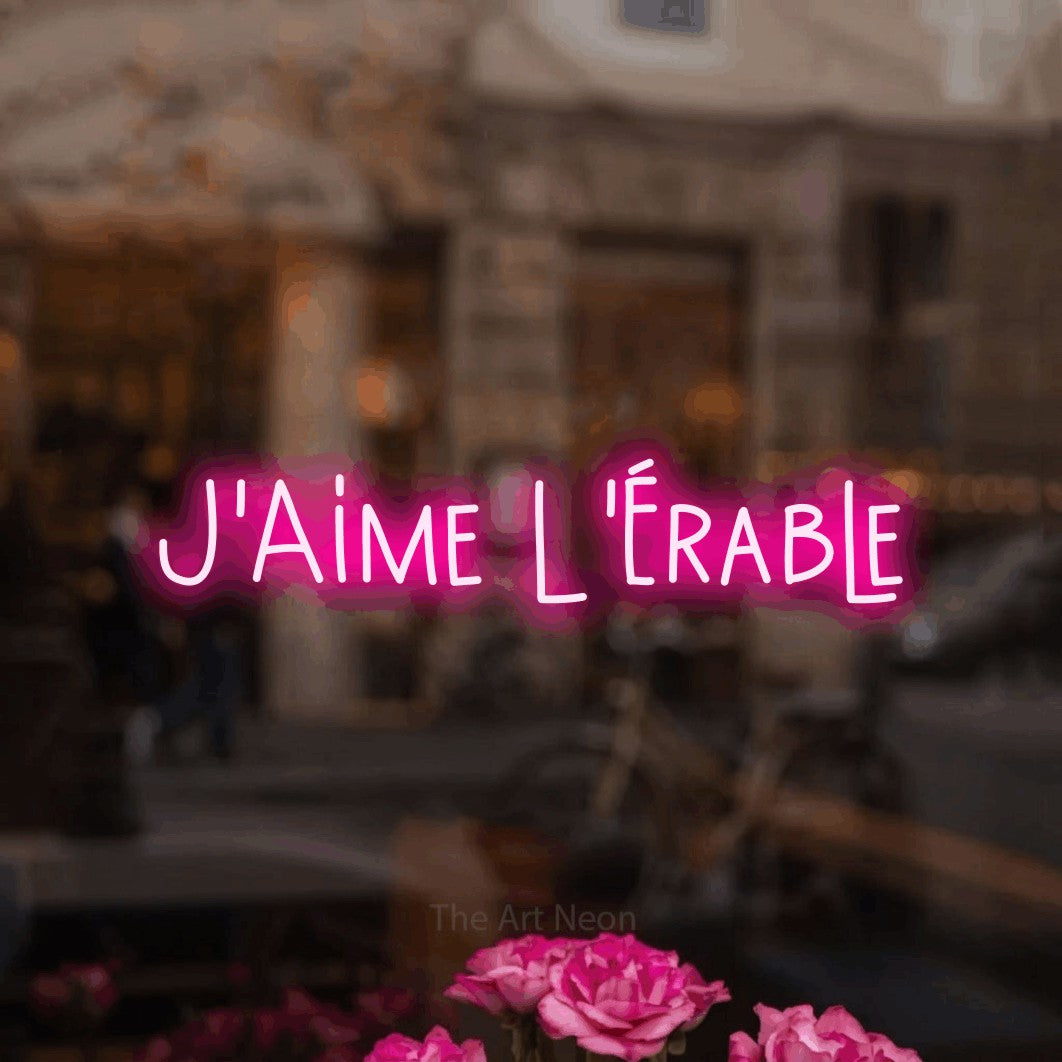 “J'aime l'érable” LED neon sign in hot pink, with narrow hand-drawn lettering, shown against a blurred café-bar interior.