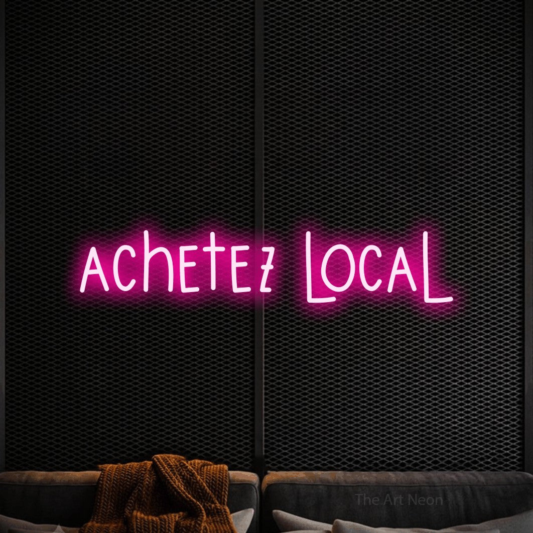 “Achetez local” LED neon sign in hot pink, with narrow hand-drawn lettering, shown against a modern dark-toned seating area.