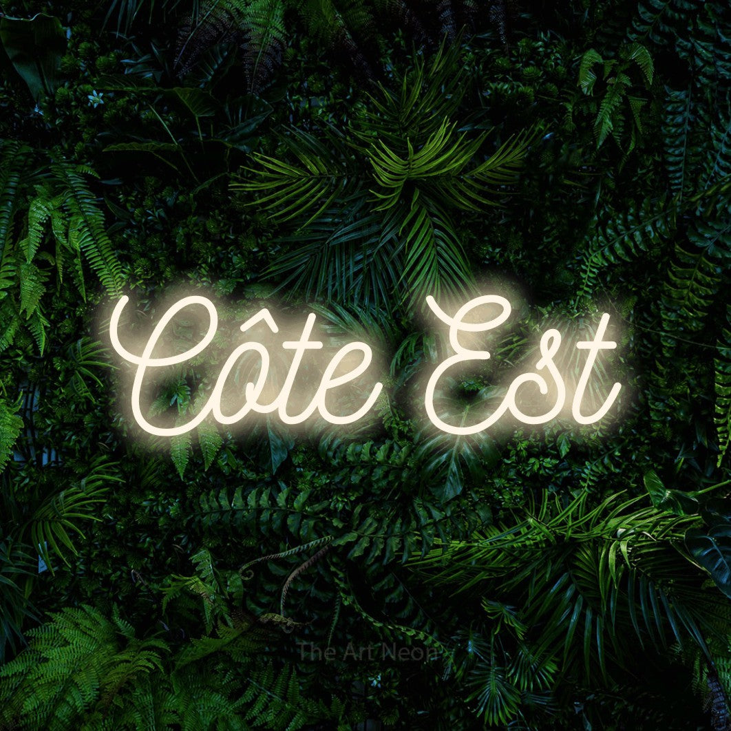 “Côte est” LED neon sign in warm white, with ornate cursive lettering, shown against dense tropical foliage.