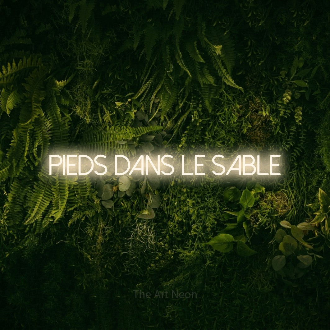 Composition en néon LED « Pieds dans le sable » en blanc chaud, en lettres fines sans empattement, mise en valeur sur un fond sombre de mousse et de forêt, pour un décor inspiré de la nature.