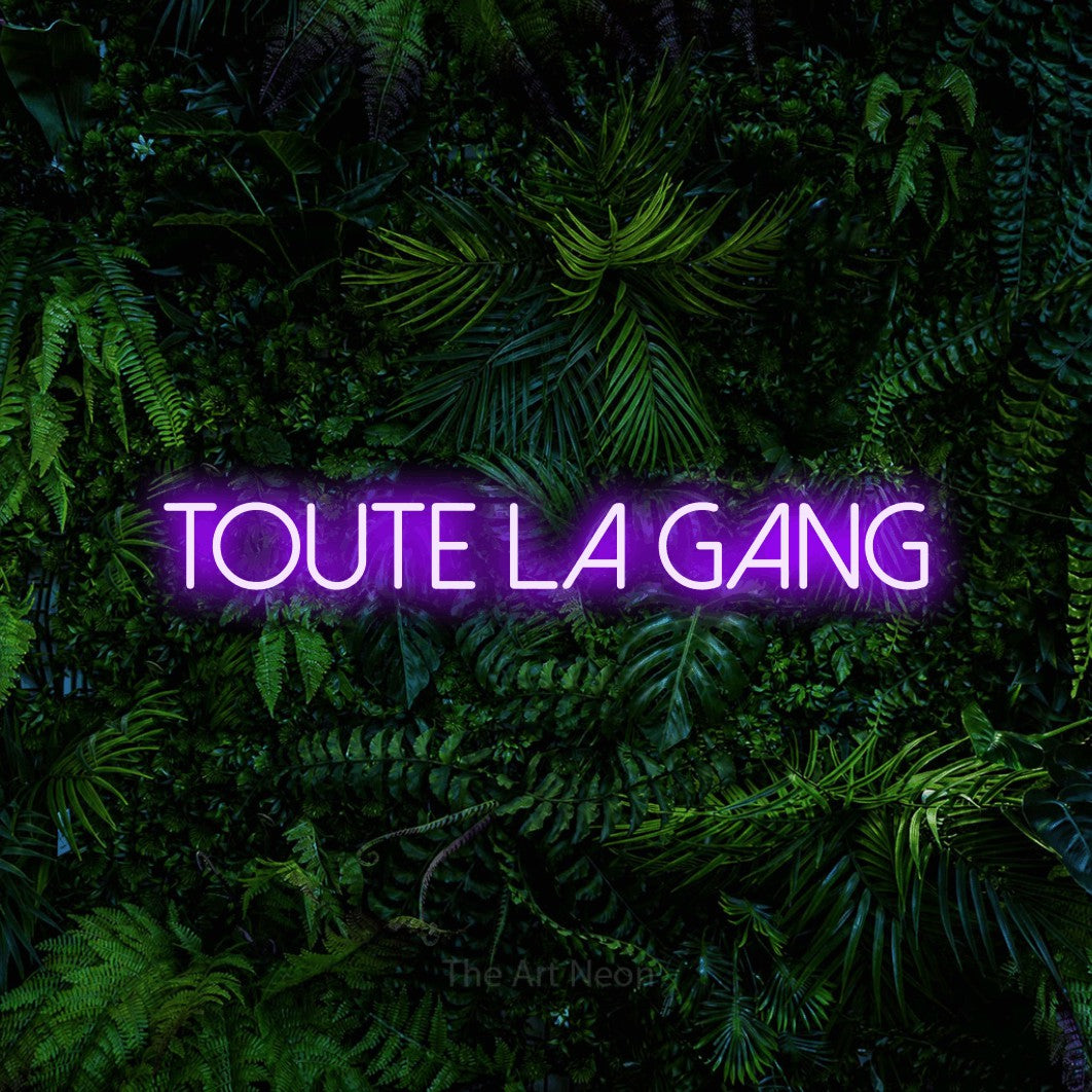 “Toute la gang” LED neon sign in purple, with thin sans-serif lettering, shown against dense tropical foliage.