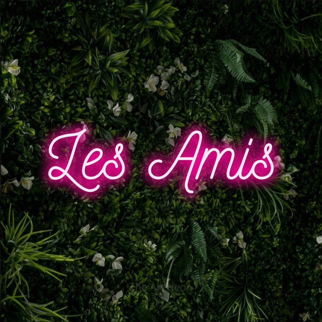 “Les amis” LED neon sign in hot pink, with ornate cursive lettering, shown against a dense green plant wall.