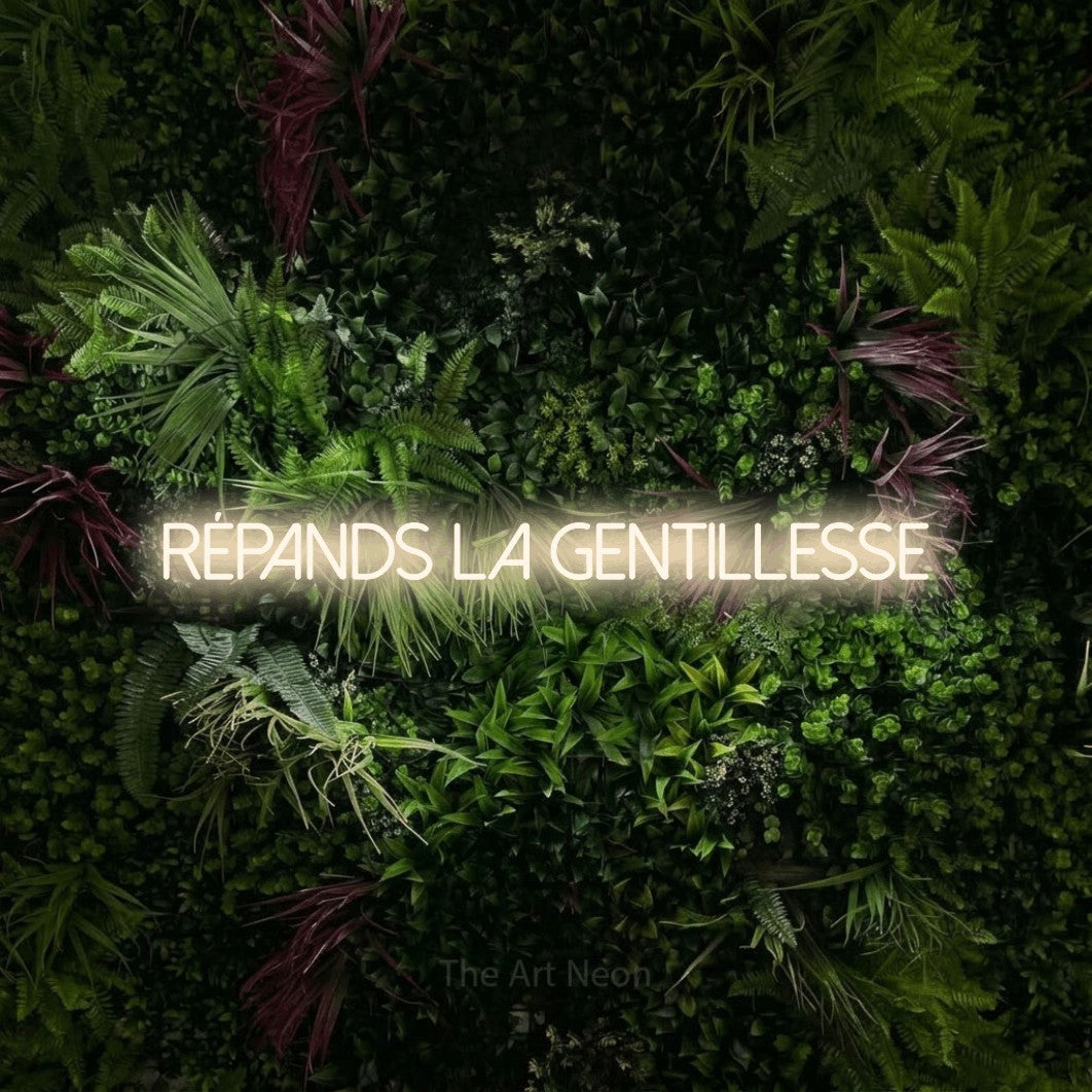“Répands la gentillesse” LED neon sign in warm white, with thin sans-serif lettering, shown against a plant wall with pink flowers.