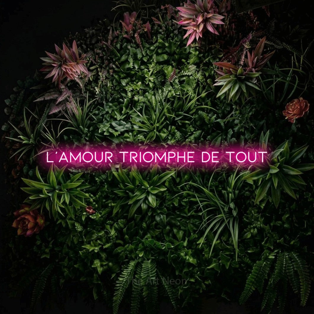 “L'amour triomphe de tout” LED neon sign in hot pink, with thin, widely spaced lettering, shown against a flowering plant wall.