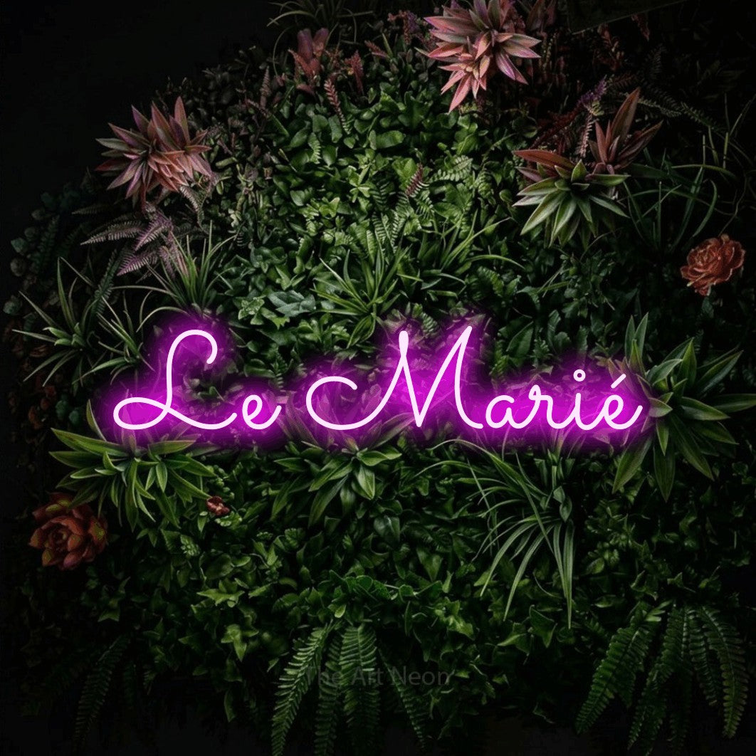 “Le marié” LED neon sign in pink, with looped cursive lettering, shown against a flowering plant wall.
