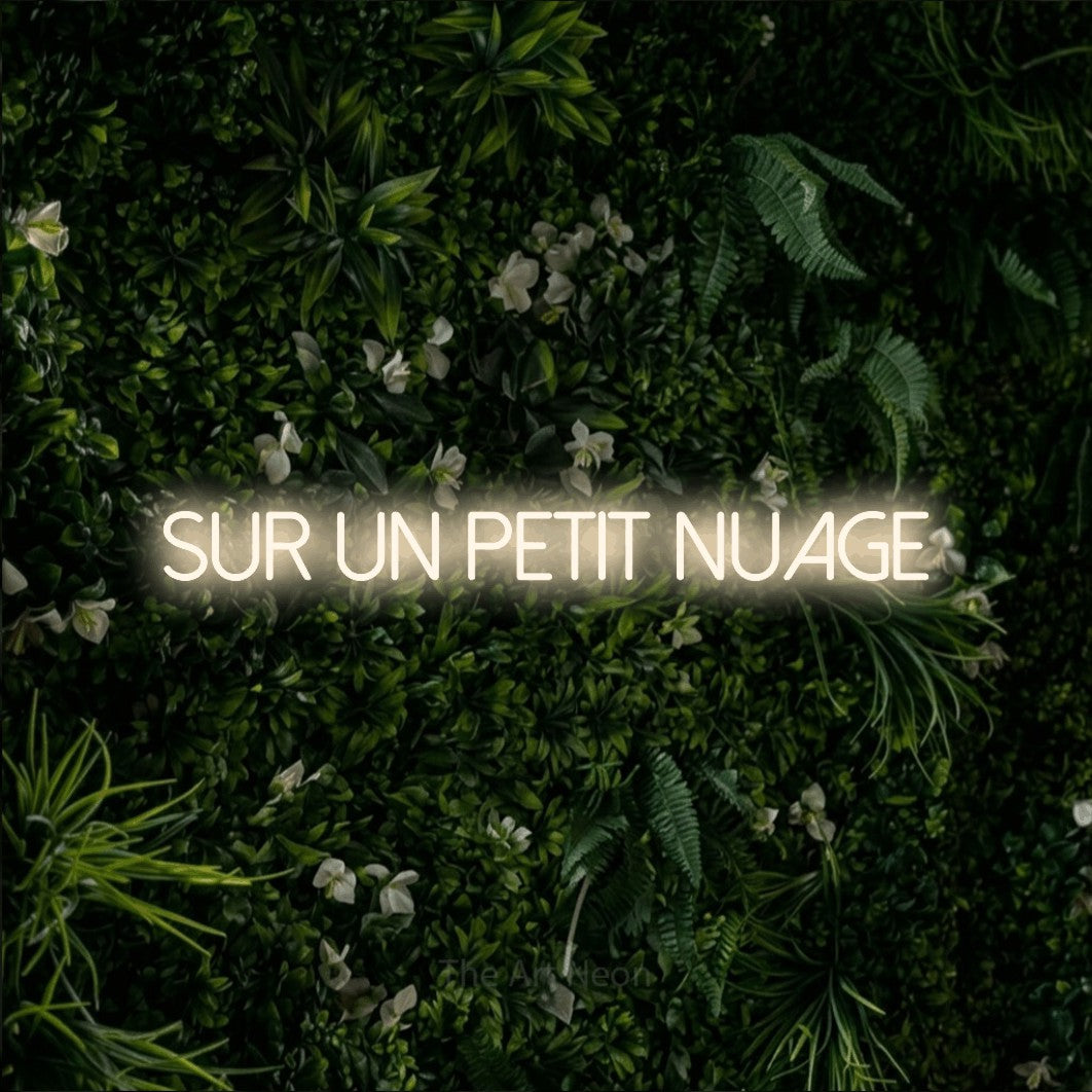 “Sur un petit nuage” LED neon sign in warm white, with thin sans-serif lettering, shown against a dense green plant wall.