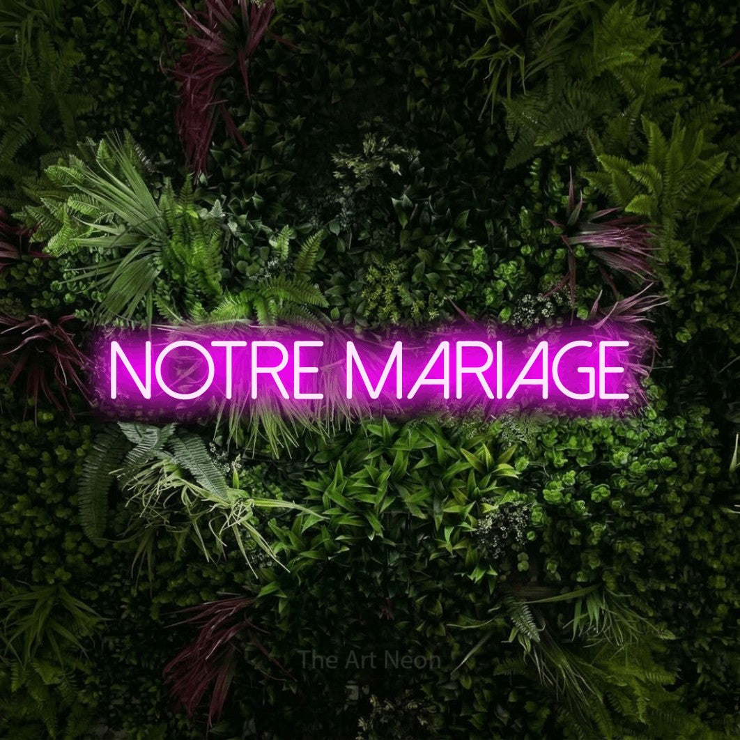 “Notre mariage” LED neon sign in pink, with thin sans-serif lettering, shown against a plant wall with pink flowers.