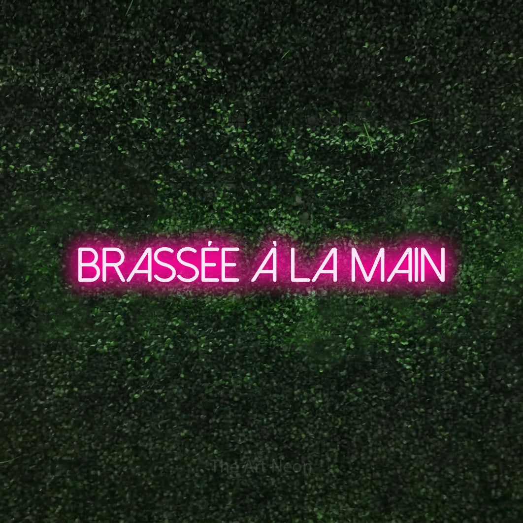 “Brassée à la main” LED neon sign in hot pink, with thin sans-serif lettering, shown against a textured green hedge.