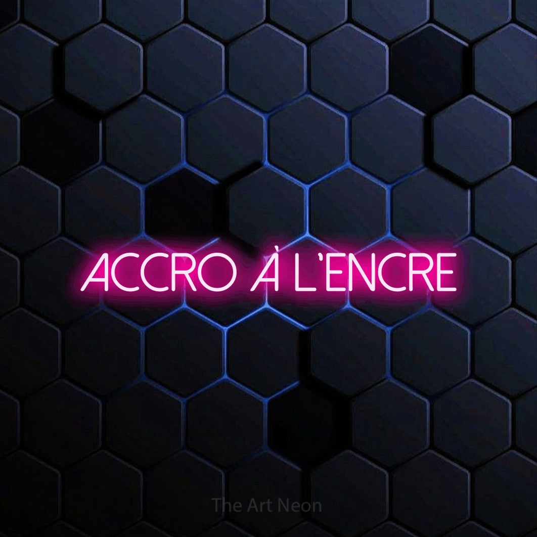 “Accro à l'encre” LED neon sign in hot pink, with thin sans-serif lettering, shown against a dark blue honeycomb pattern.