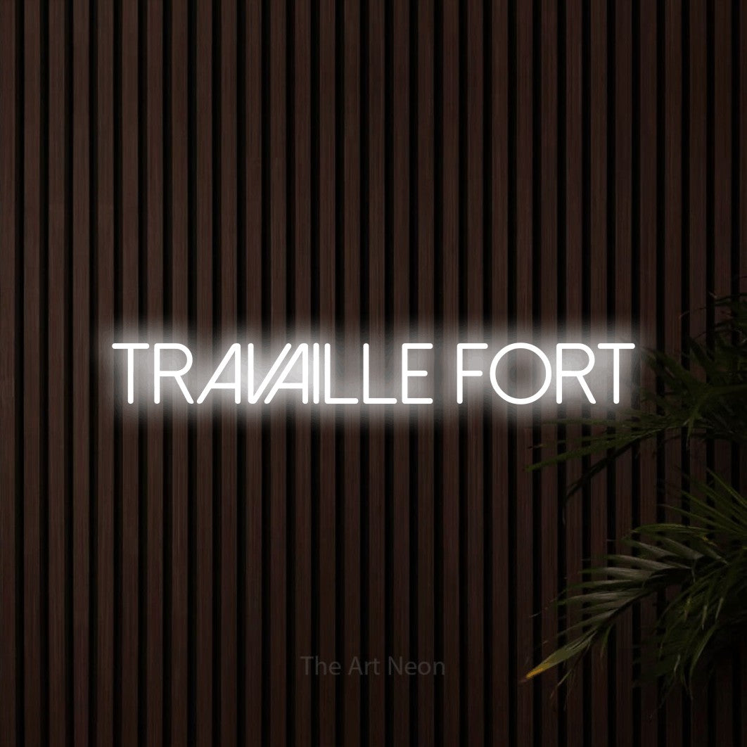 “Travaille fort” LED neon sign in cool white, with thin sans-serif lettering, shown against dark wooden wall slats.