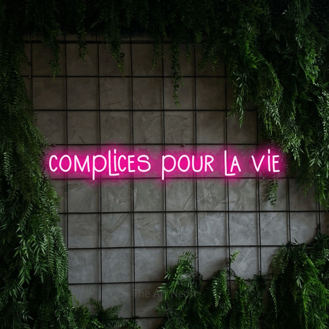 “Complices pour la vie” LED neon sign in hot pink, with narrow hand-drawn lettering, shown against an ivy-covered concrete grid.