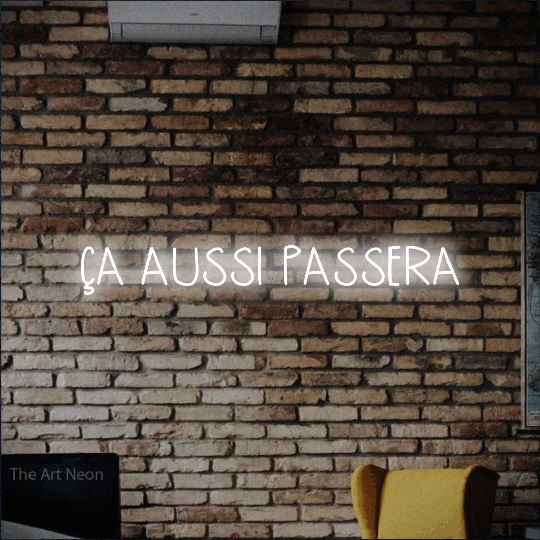 “Ça aussi passera” LED neon sign in cool white, with narrow hand-drawn lettering, shown against warm-toned brickwork.