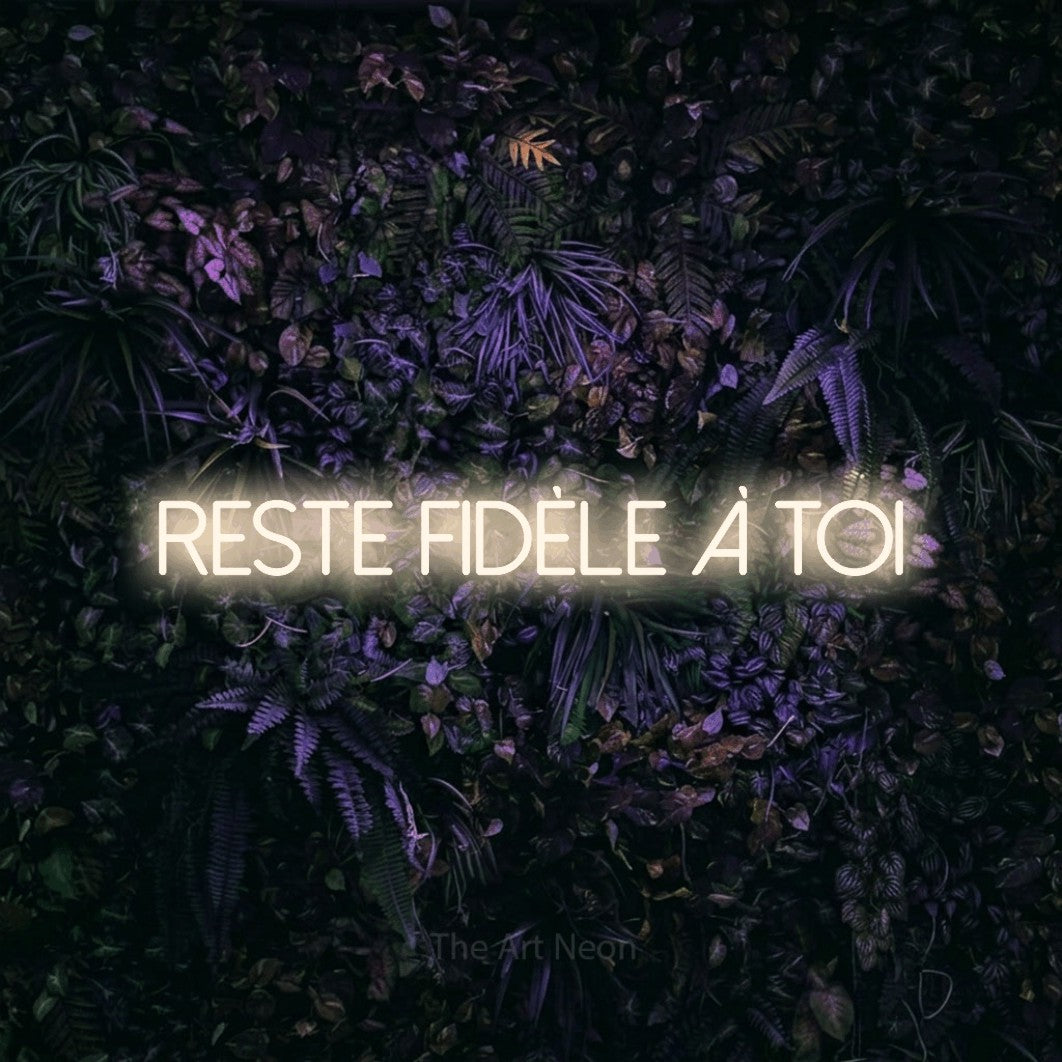 “Reste fidèle à toi” LED neon sign in warm white, with thin sans-serif lettering, shown against a dark wall with purple flowers.