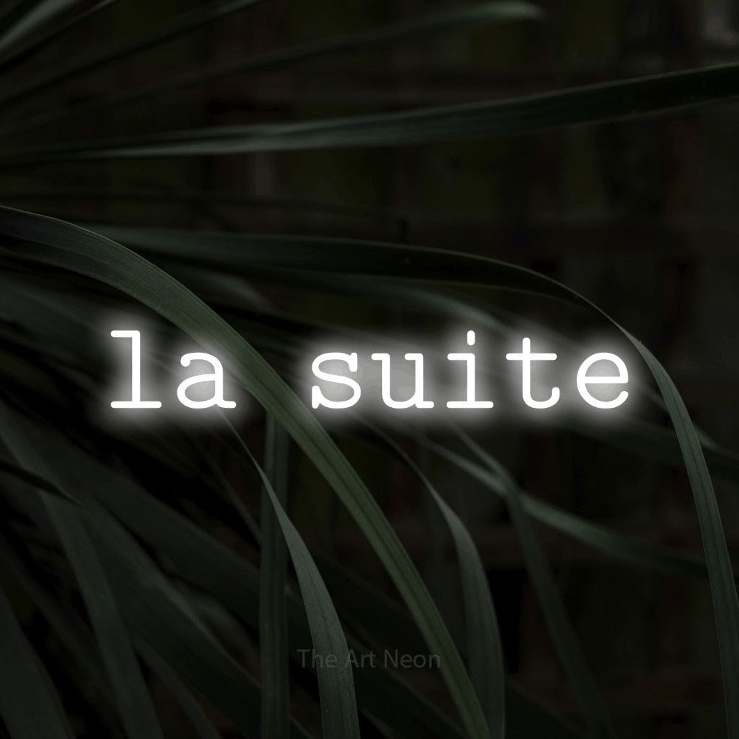 Néon LED « La suite » en blanc froid, en caractères de machine à écrire, mis en scène sur un feuillage sombre en gros plan, pour un hôtel.