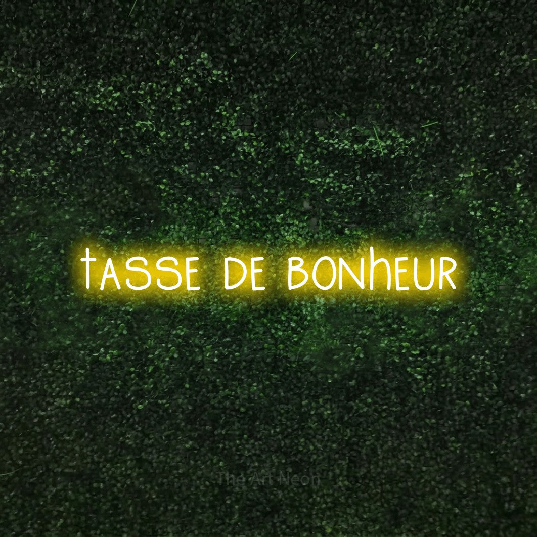“Tasse de bonheur” LED neon sign in yellow, with narrow hand-drawn lettering, shown against a textured green hedge.