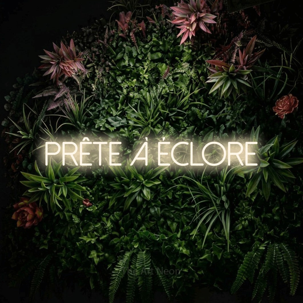 “Prête à éclore” LED neon sign in warm white, with thin sans-serif lettering, shown against a flowering plant wall.