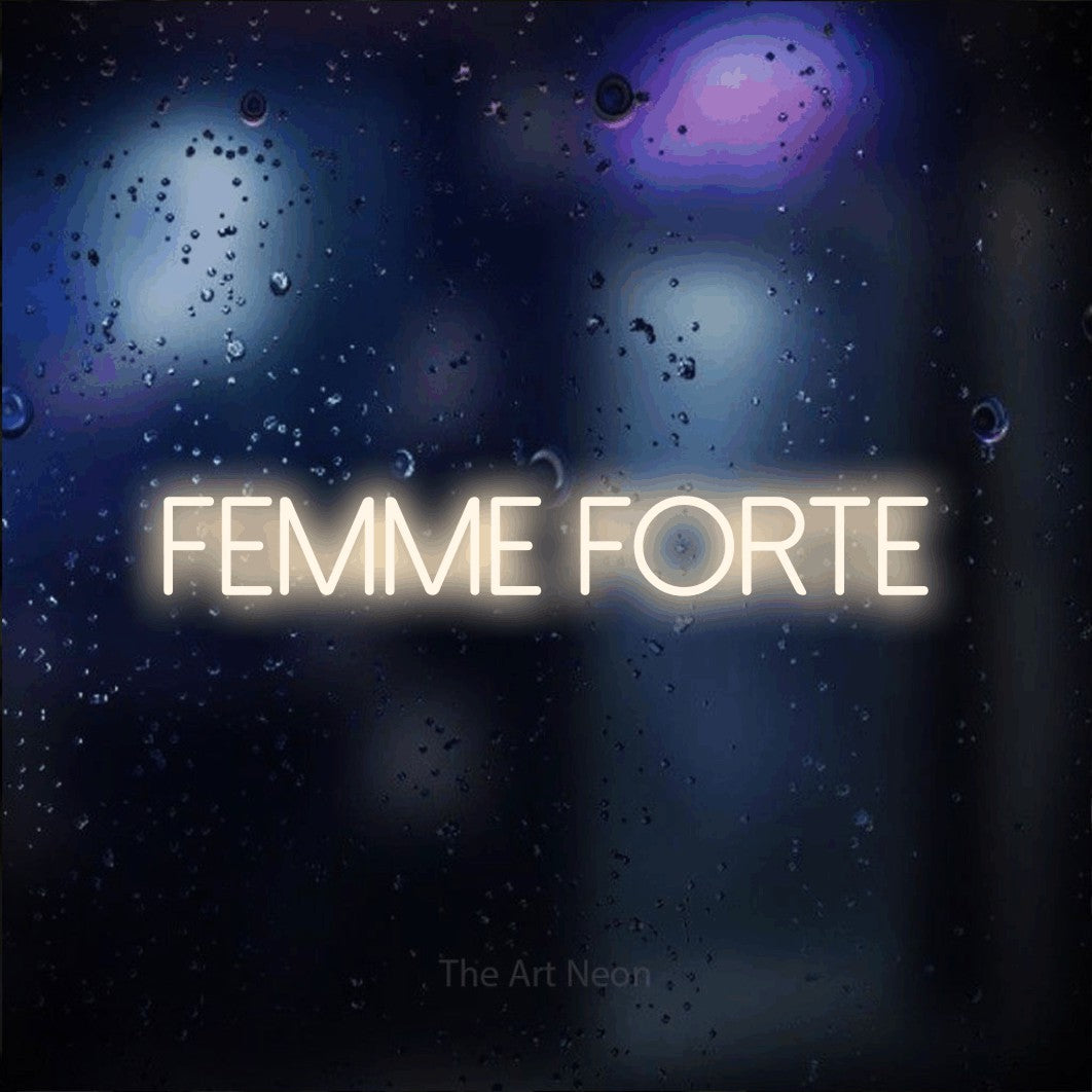 “Femme forte” LED neon sign in warm white, with thin sans-serif lettering, shown against glass with blue and purple reflections.