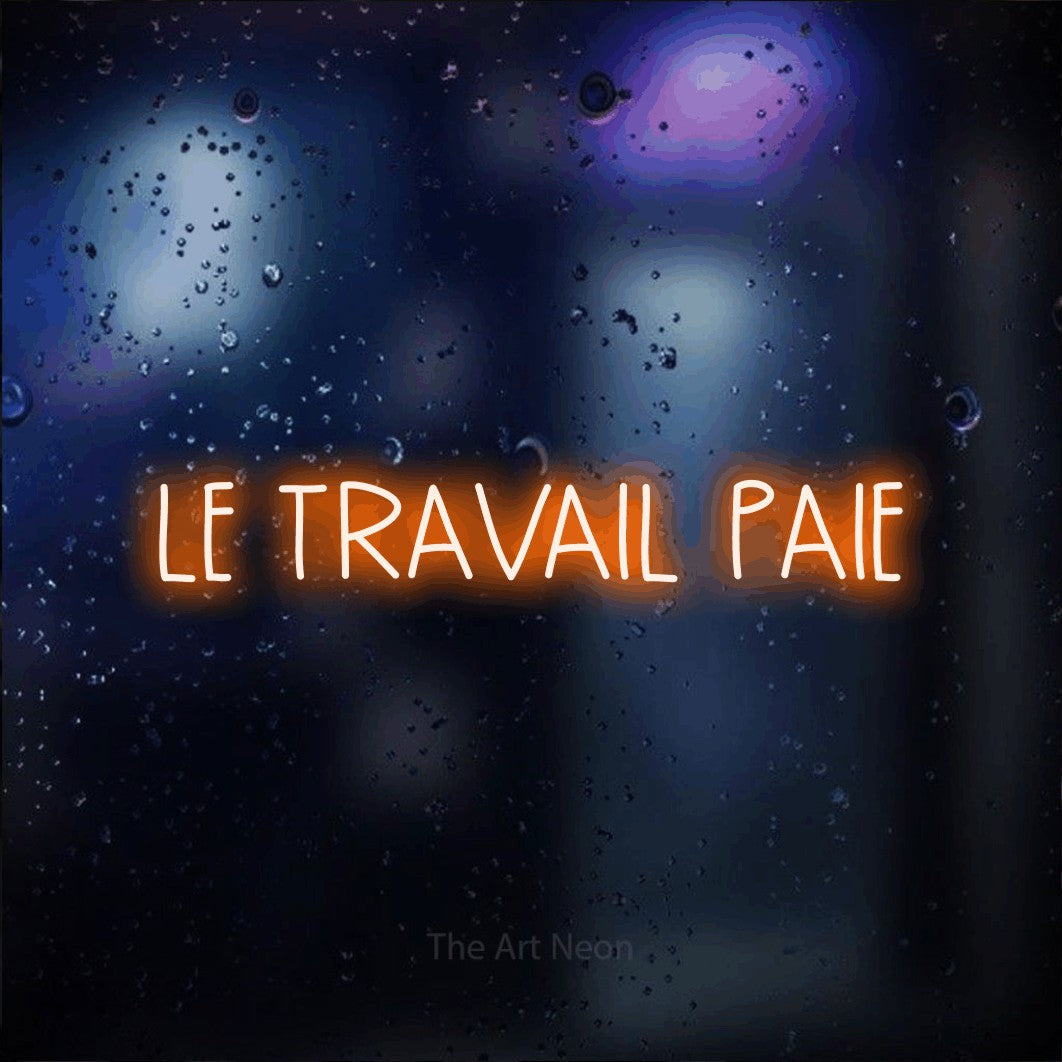 “Le travail paie” LED neon sign in orange, with narrow hand-drawn lettering, shown against glass with blue and purple reflections.