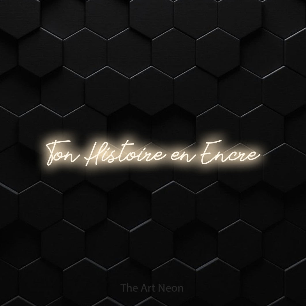 “Ton histoire en encre” LED neon sign in warm white, with cursive lettering, shown against black hexagonal wall tiles.