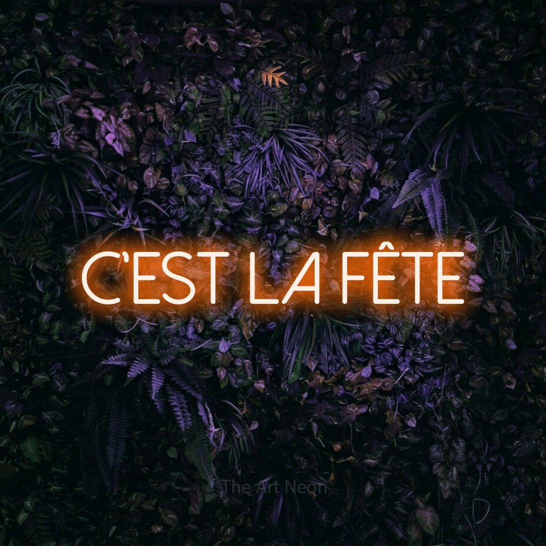 Décoration murale en néon LED « C'est la fête » en orange, en lettres fines sans empattement, installée sur un mur sombre aux fleurs violettes, pour une fête.