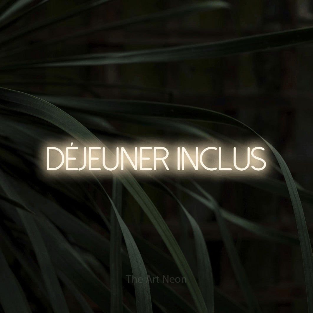 “Déjeuner inclus” LED neon sign in warm white, with thin sans-serif lettering, shown against a close-up of dark foliage.